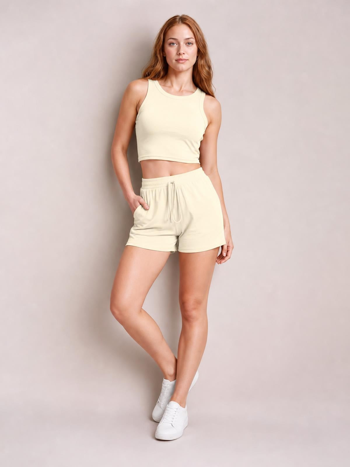 Model poses in a cream round neck tank and drawstring shorts set with a cropped fitted silhouette, elastic waistband, and side pockets.
