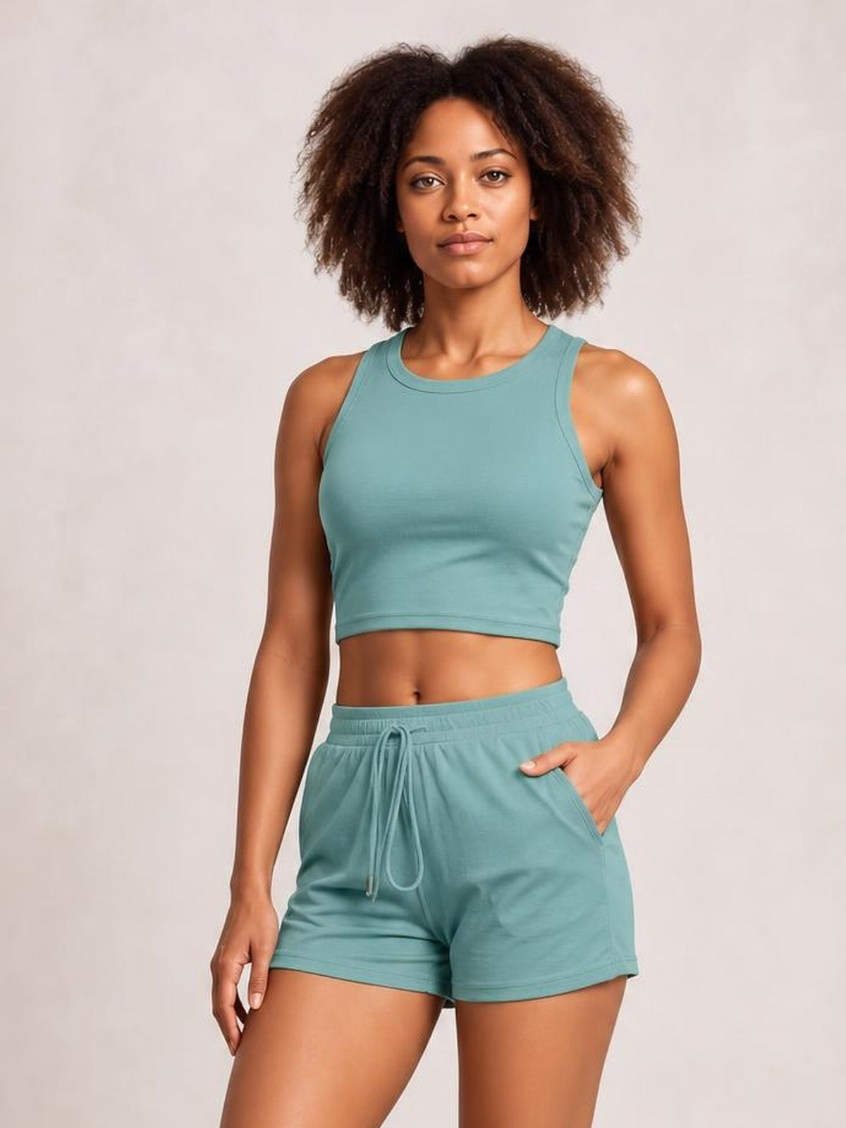 Model wearing a teal round neck tank and drawstring shorts set, featuring a fitted cropped silhouette, relaxed shorts, side pockets, and one hand in a pocket.