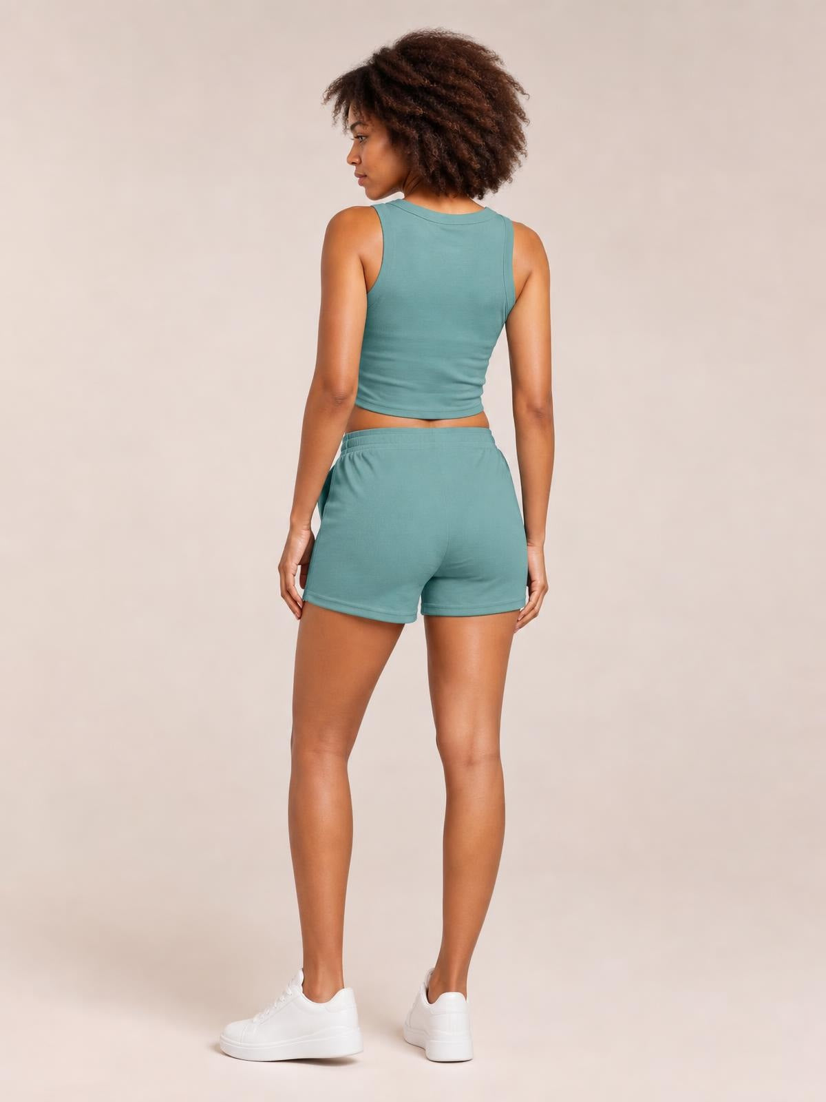 A model stands turned away in a teal round-neck tank with a fitted cropped silhouette and matching relaxed drawstring shorts with an elastic waistband.