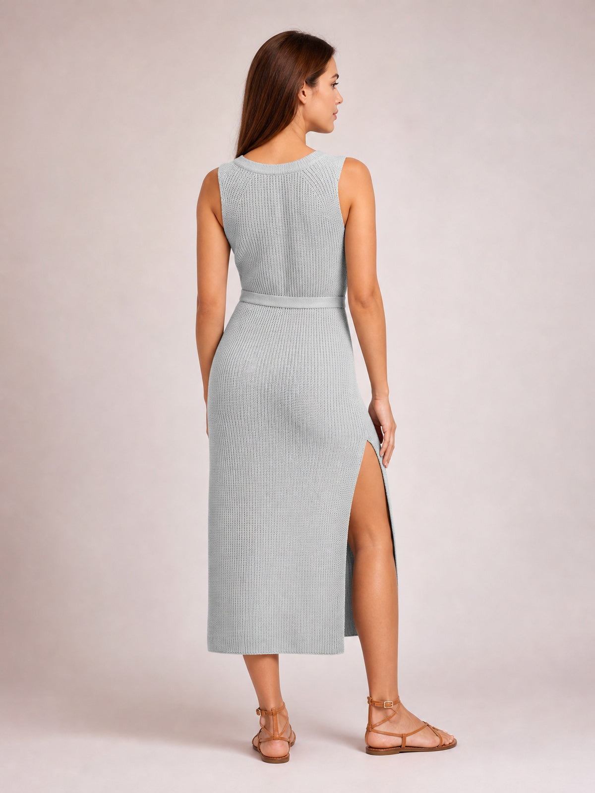 A model poses from the back in a solid light gray knit midi dress with a round neck, sleeveless cut, waist tie belt, streamlined silhouette, and side slit.