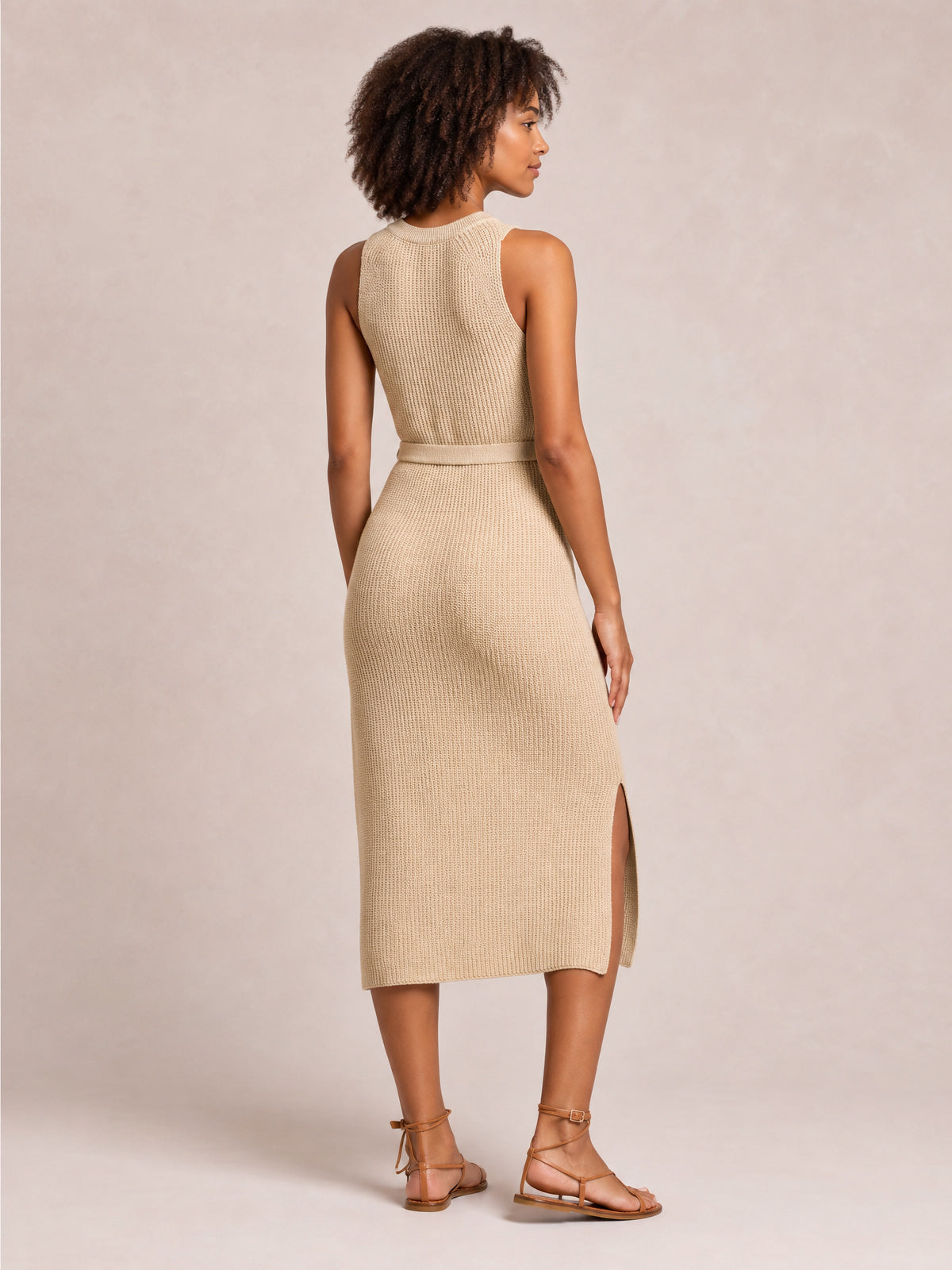 Model looks over her shoulder in a solid beige sleeveless knit sweater dress with a round neck, tie belt, streamlined midi silhouette, and side slit.