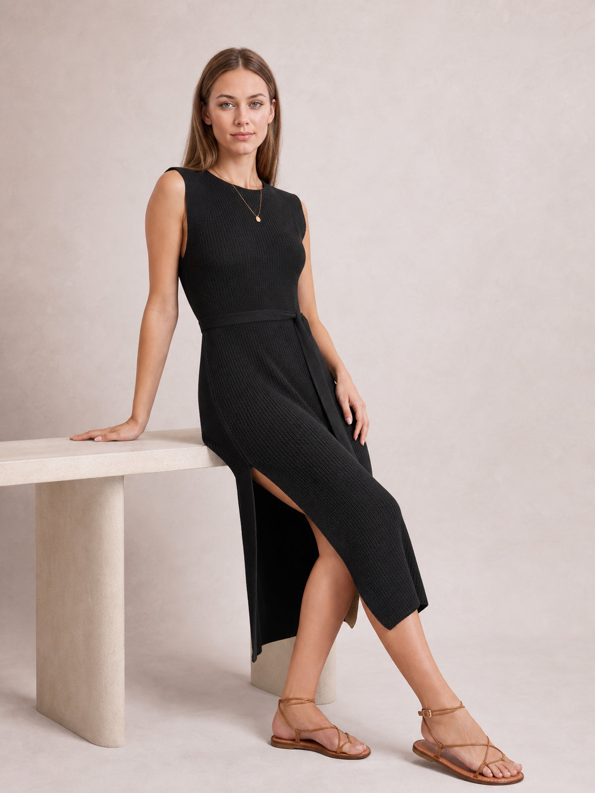 Model seated in a black sleeveless sweater dress with a round neck, tie belt, stretchy knit texture, streamlined midi silhouette, and dramatic side slit.