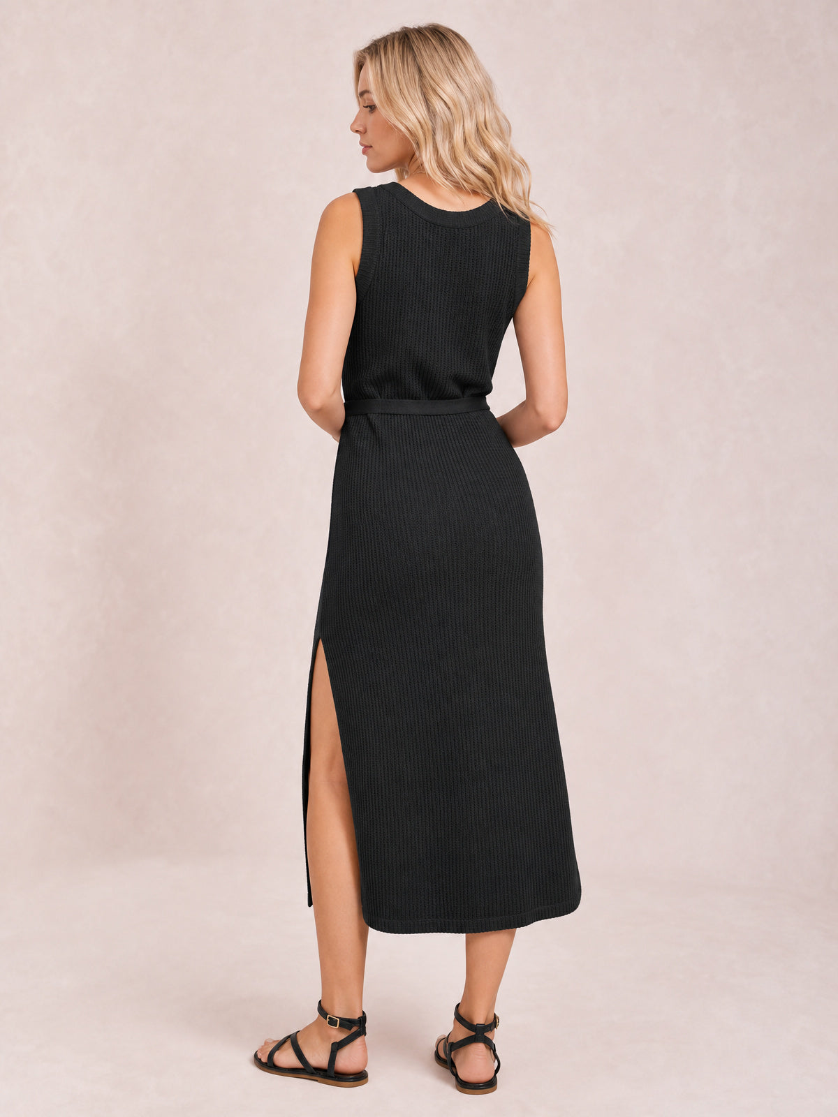 A model turned away wears a black sleeveless sweater midi dress with a round neckline, tie waist belt, fitted silhouette, and high side slit.