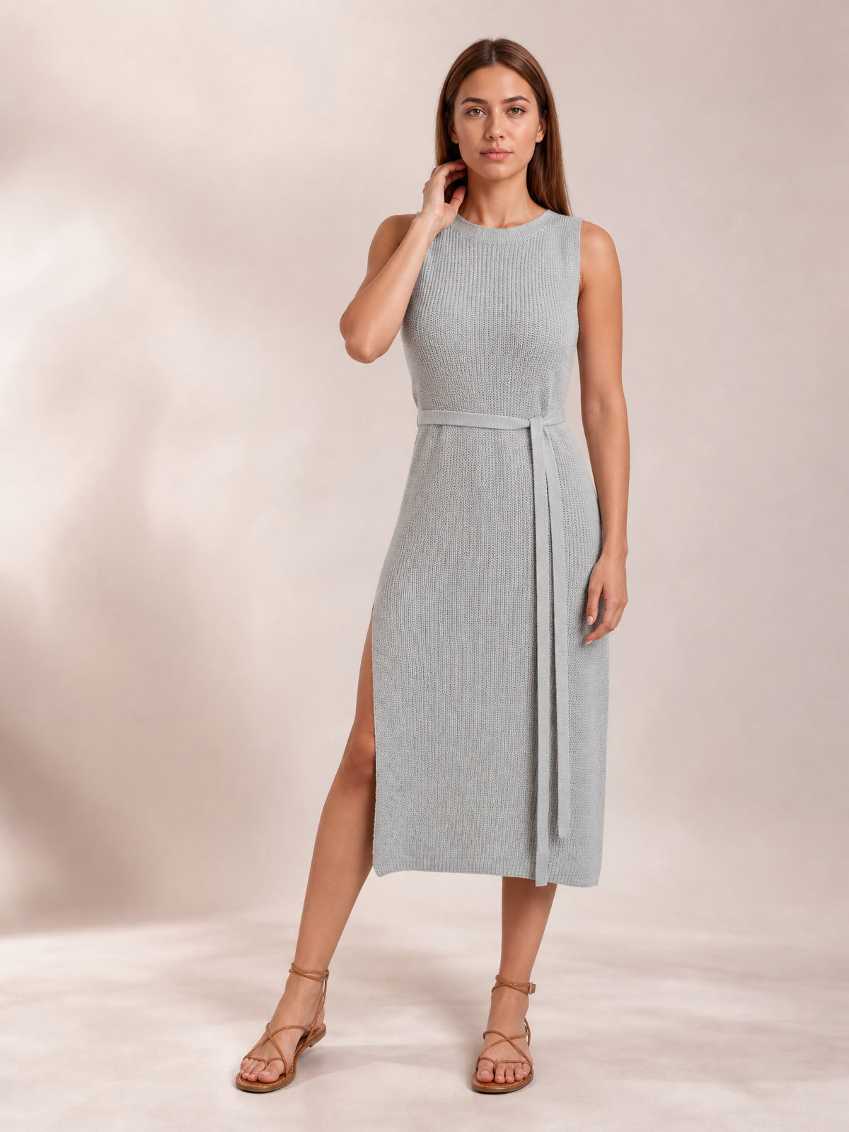 A model poses in a light gray sleeveless sweater dress with a round neck, fitted midi silhouette, tie belt, and side slit.