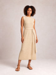 Model stands in a beige sleeveless sweater dress with a round neck, tie belt, textured knit, streamlined midi silhouette, and subtle side slit.