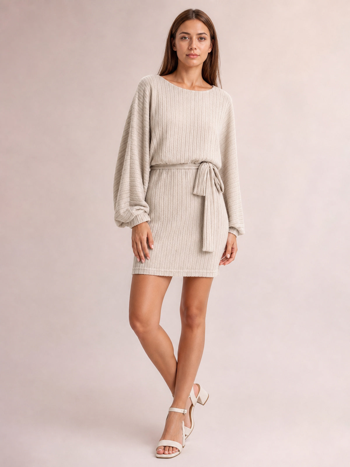 A model poses with crossed legs in a khaki ribbed mini dress featuring a round neck, relaxed long sleeves, tie waist, and straight silhouette.