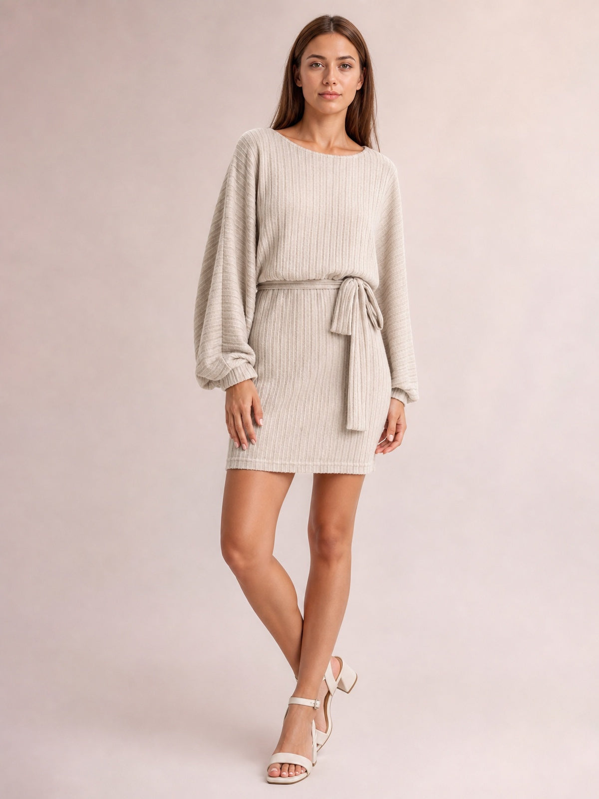 A model poses with crossed legs in a khaki ribbed mini dress featuring a round neck, relaxed long sleeves, tie waist, and straight silhouette.