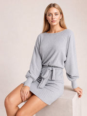 Model seated in a gray textured mini dress with a round neck, balloon sleeves, fitted cuffs, and a self-tie waist creating a defined silhouette.