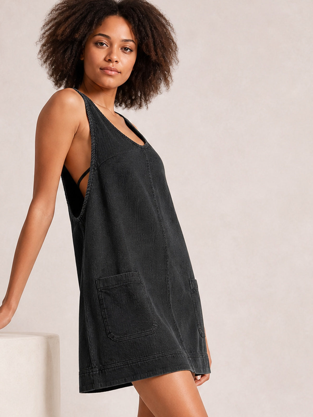 A model poses in a black round-neck mini dress with wide shoulder straps, a relaxed A-line silhouette, front patch pockets, and a structured hem.