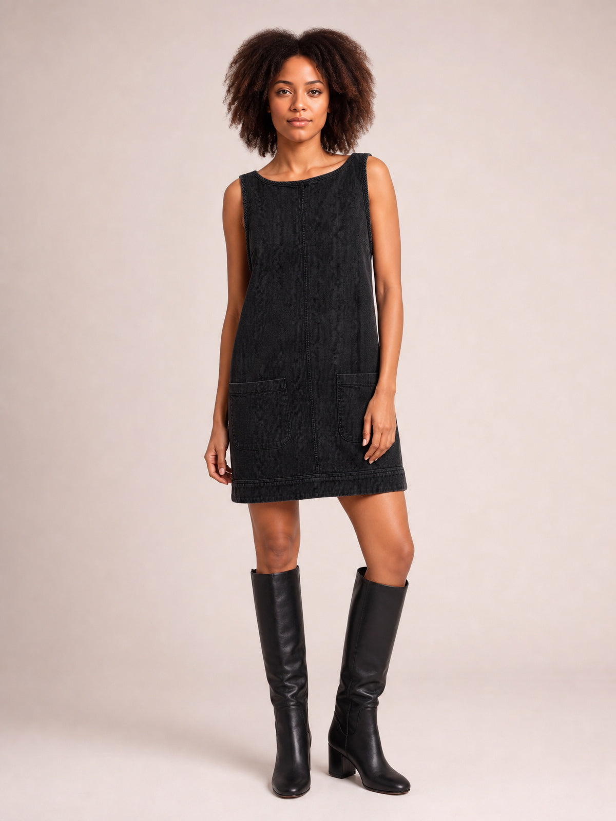 Model poses in a black round-neck mini dress with wide straps, an A-line silhouette, front patch pockets, and visible center and hem seams.