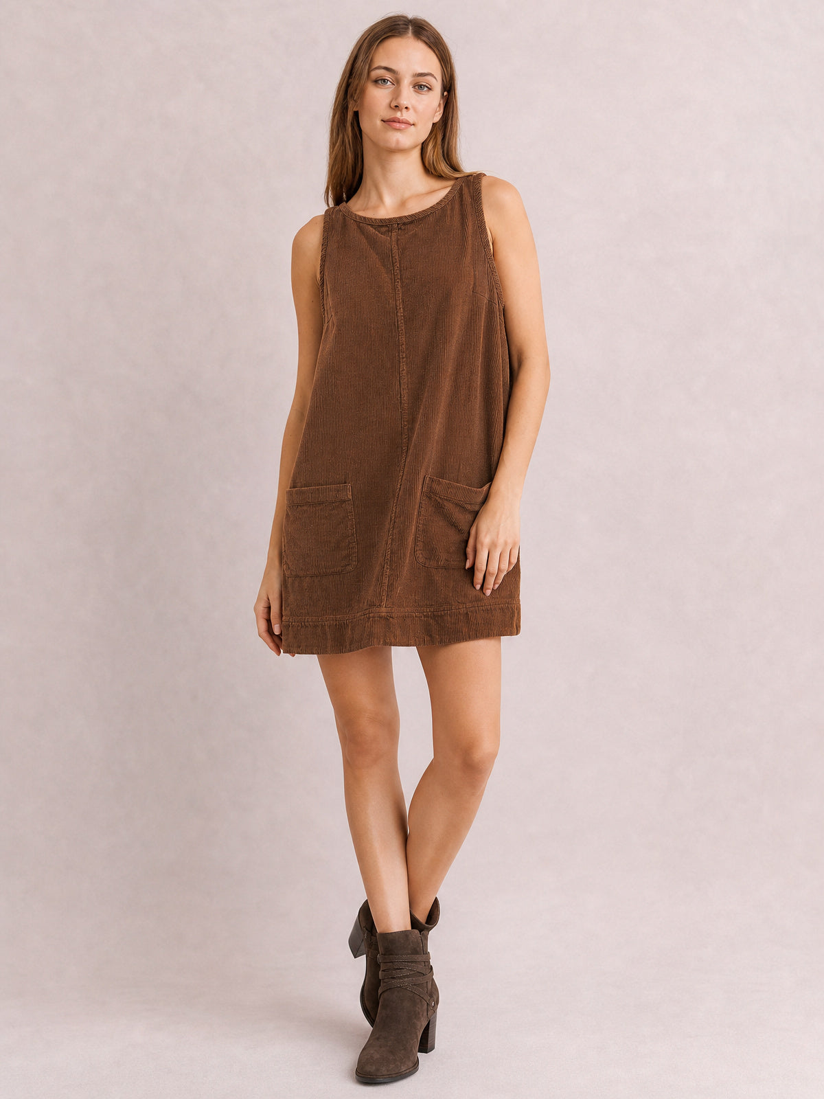 Model wears a brown round-neck mini dress with wide straps, a relaxed A-line silhouette, visible center seam, and two front patch pockets, posing with crossed legs.