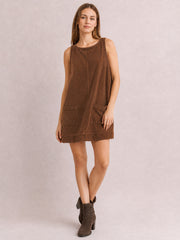 Model wears a brown round-neck mini dress with wide straps, a relaxed A-line silhouette, visible center seam, and two front patch pockets, posing with crossed legs.