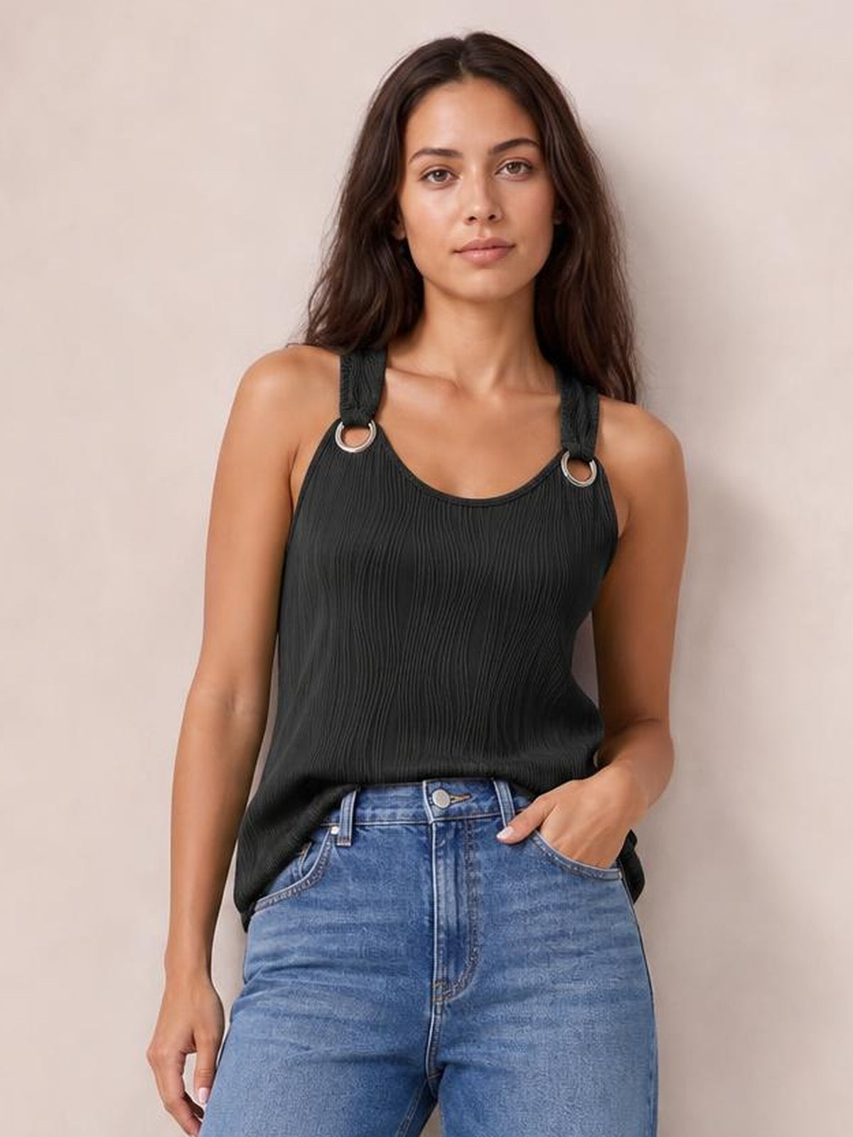 A model wears a black round neck wide strap tank with a ribbed texture, relaxed fit, and ring-accented straps, posing with one hand in her jeans pocket.