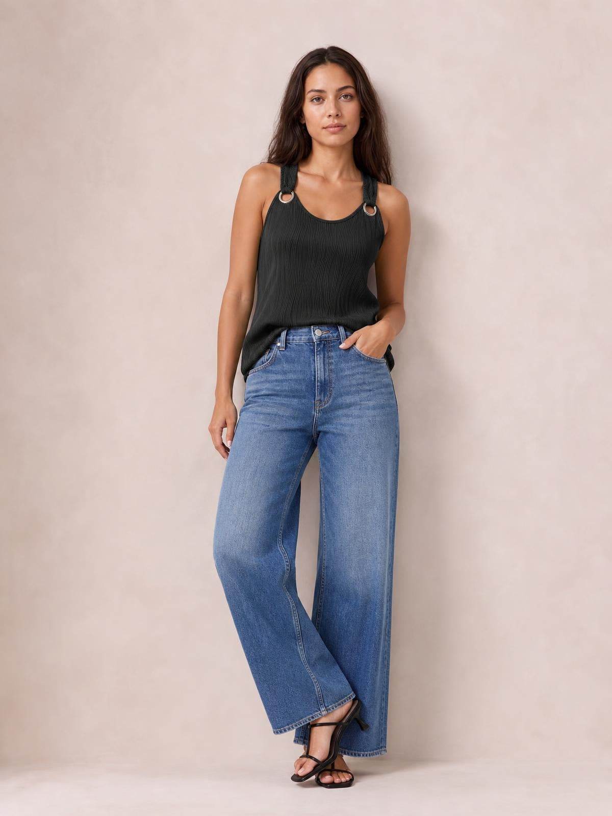 A model stands with one leg crossed, wearing a black round neck wide strap tank with a softly pleated texture and relaxed silhouette, paired with blue wide-leg jeans.