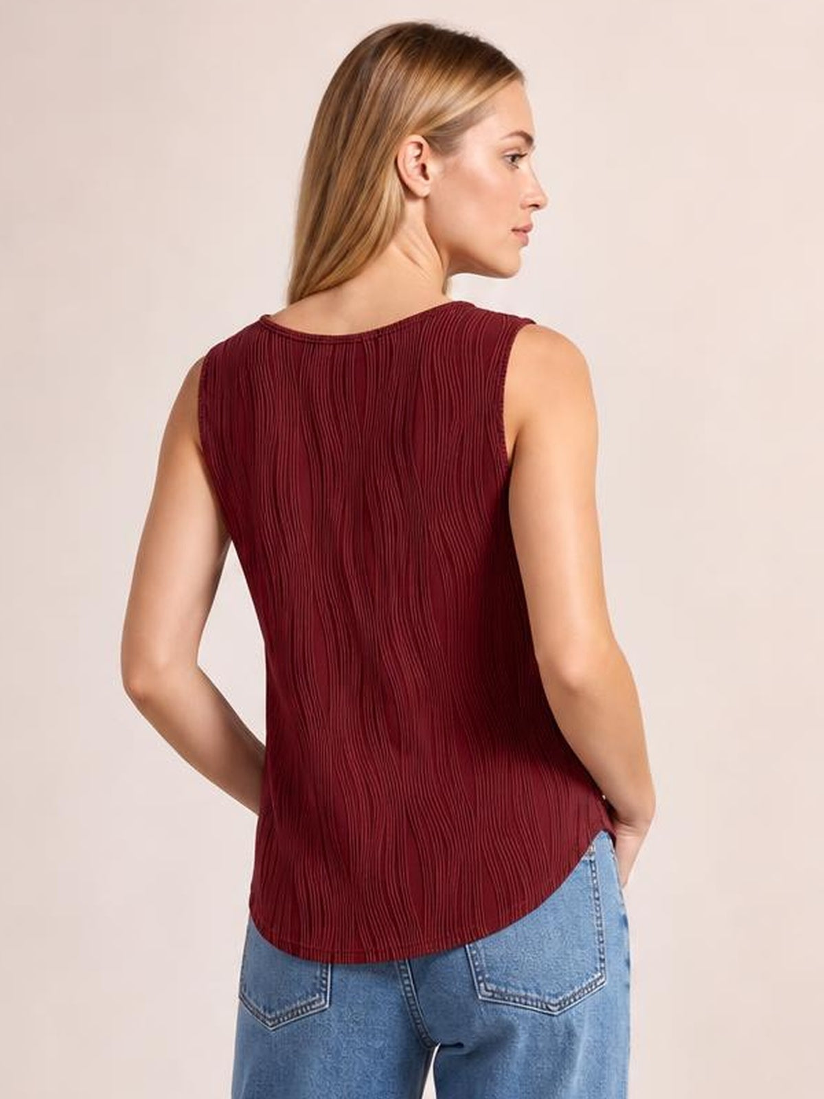 A model shows the back of a burgundy round neck wide strap tank with vertical pleated texture, a relaxed silhouette, and a curved hem.