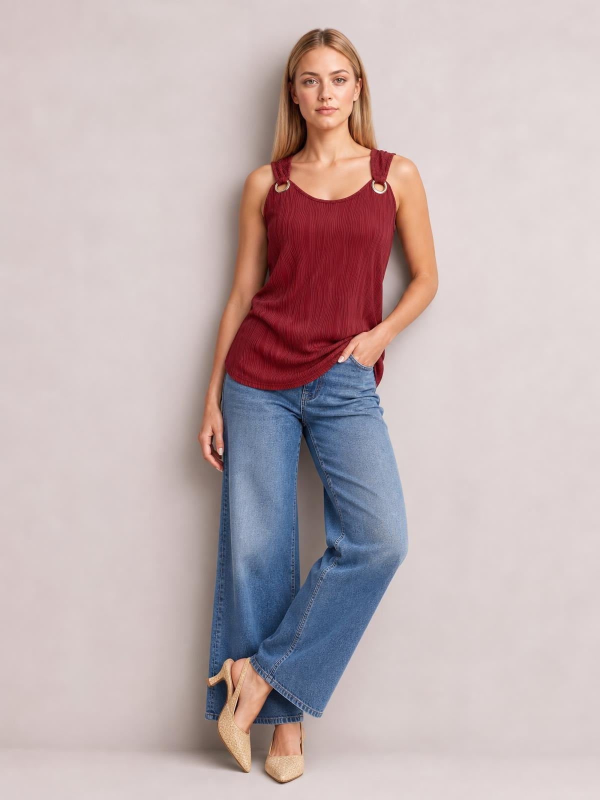 Model wears a burgundy round neck wide strap tank with a softly textured, relaxed silhouette and ring accents, posing with one hand in her jeans pocket.