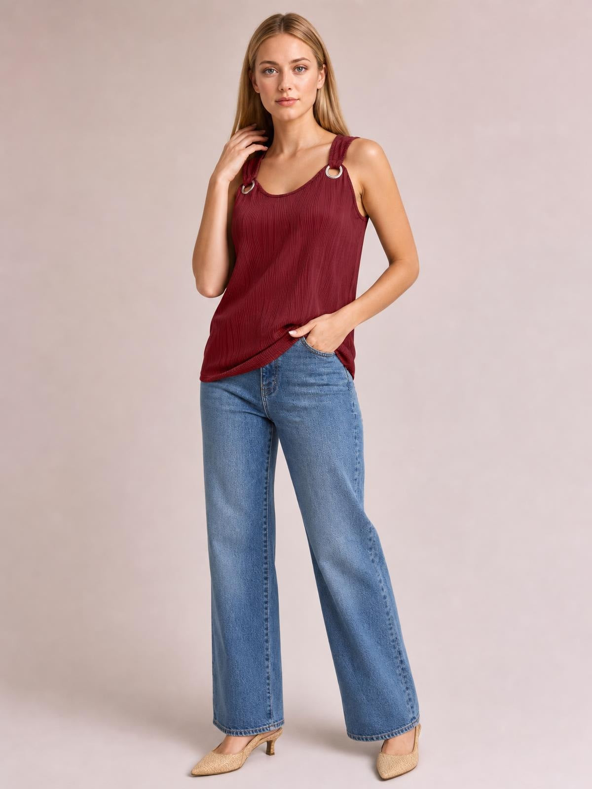 Model wears a burgundy round neck wide strap tank with a relaxed silhouette, pleated texture, and ring-accented straps, paired with wide-leg jeans.