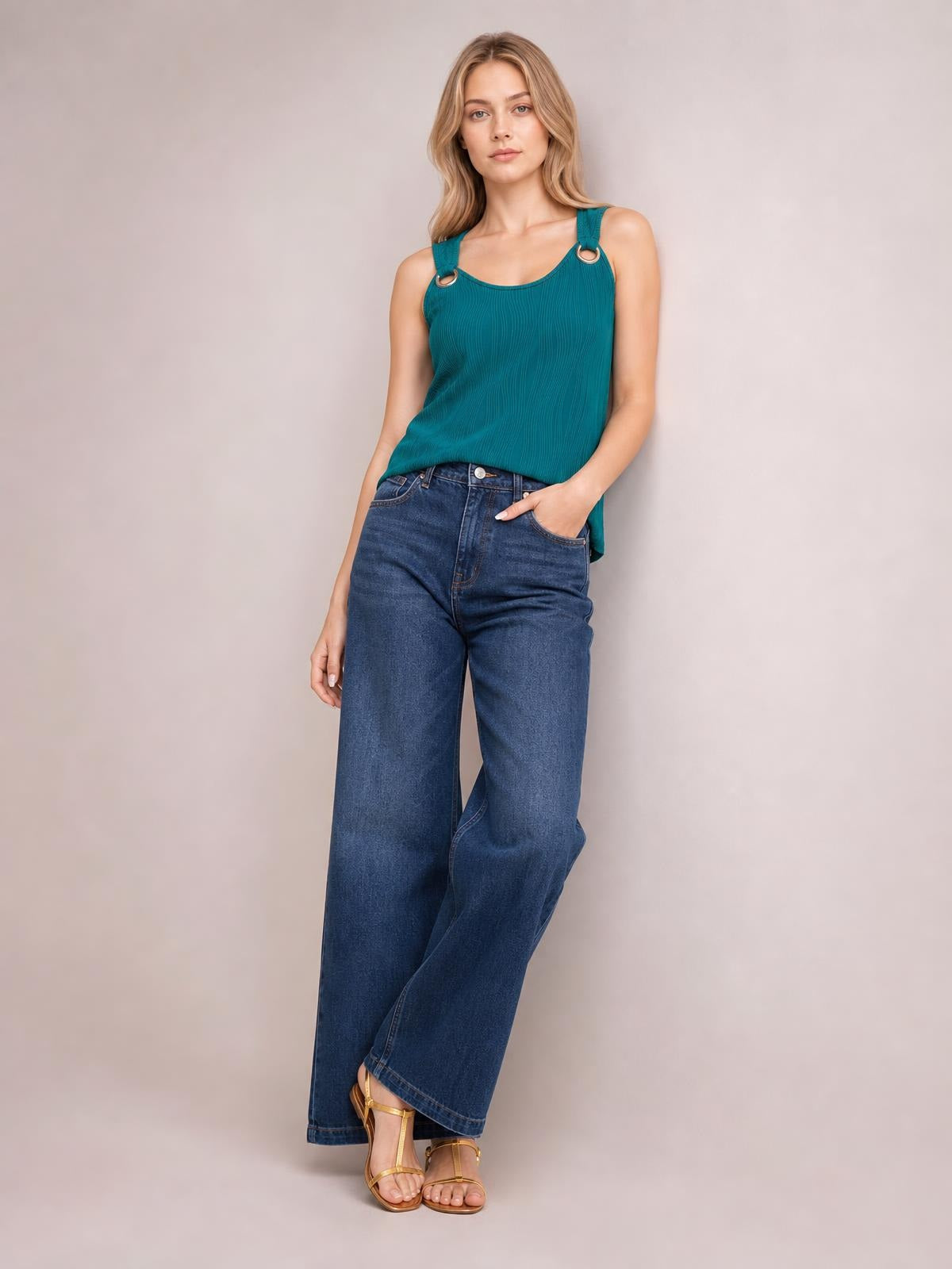 Model wears a teal round neck wide strap tank with a relaxed straight silhouette and textured finish, posing with one hand in her jeans pocket.
