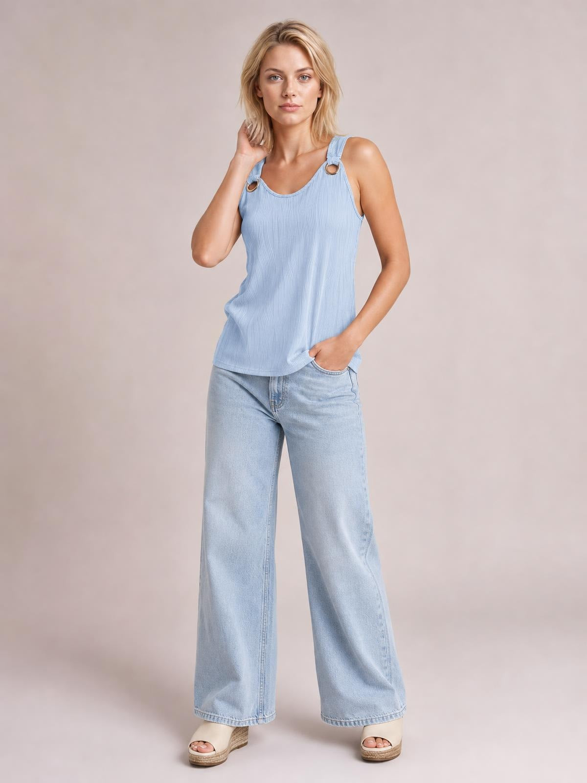 A model wears a light blue round neck wide strap tank with a relaxed straight silhouette, textured construction, and ring-accented straps, posing with one hand at her neck.