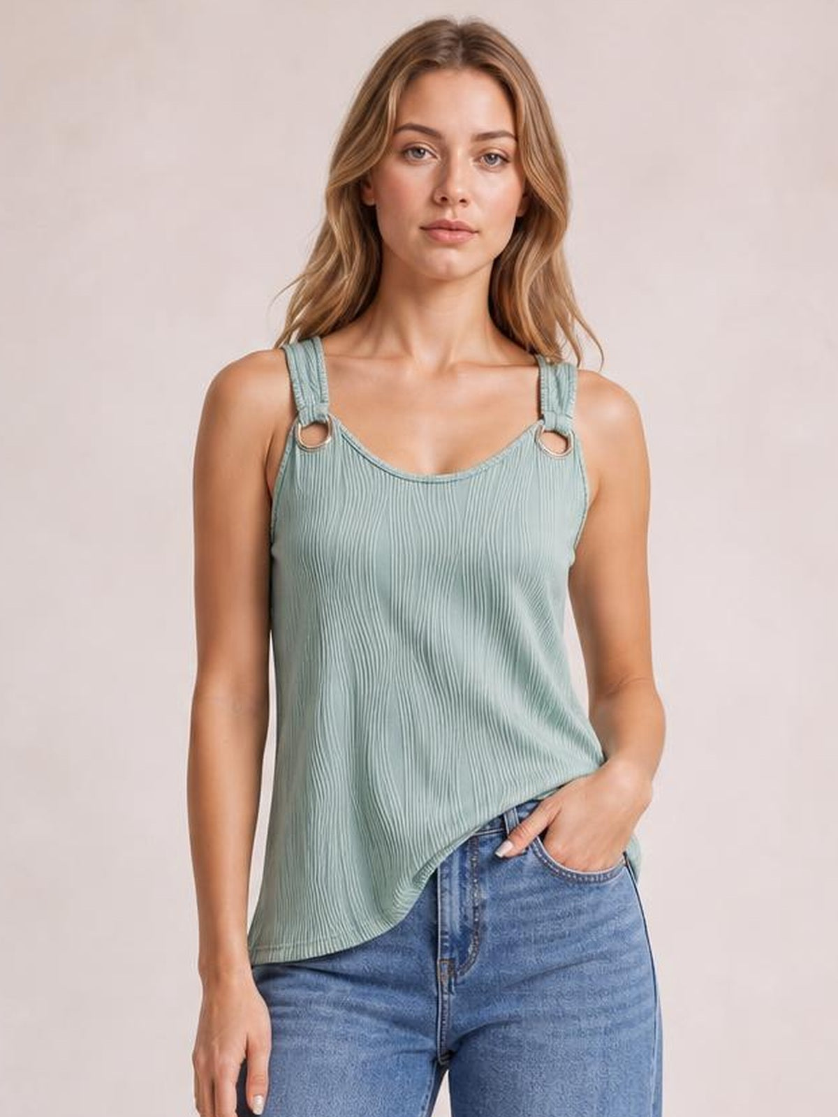 A model wears a sage green round neck wide strap tank with a ribbed texture, relaxed silhouette, ring accents, and one hand tucked into her jeans pocket.