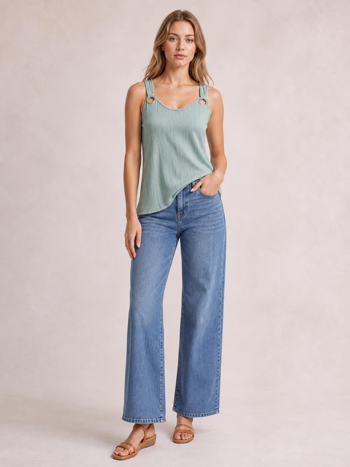 A model stands in a sage green round-neck wide-strap tank with a softly draped, ribbed silhouette and ring-accented straps, paired with jeans.