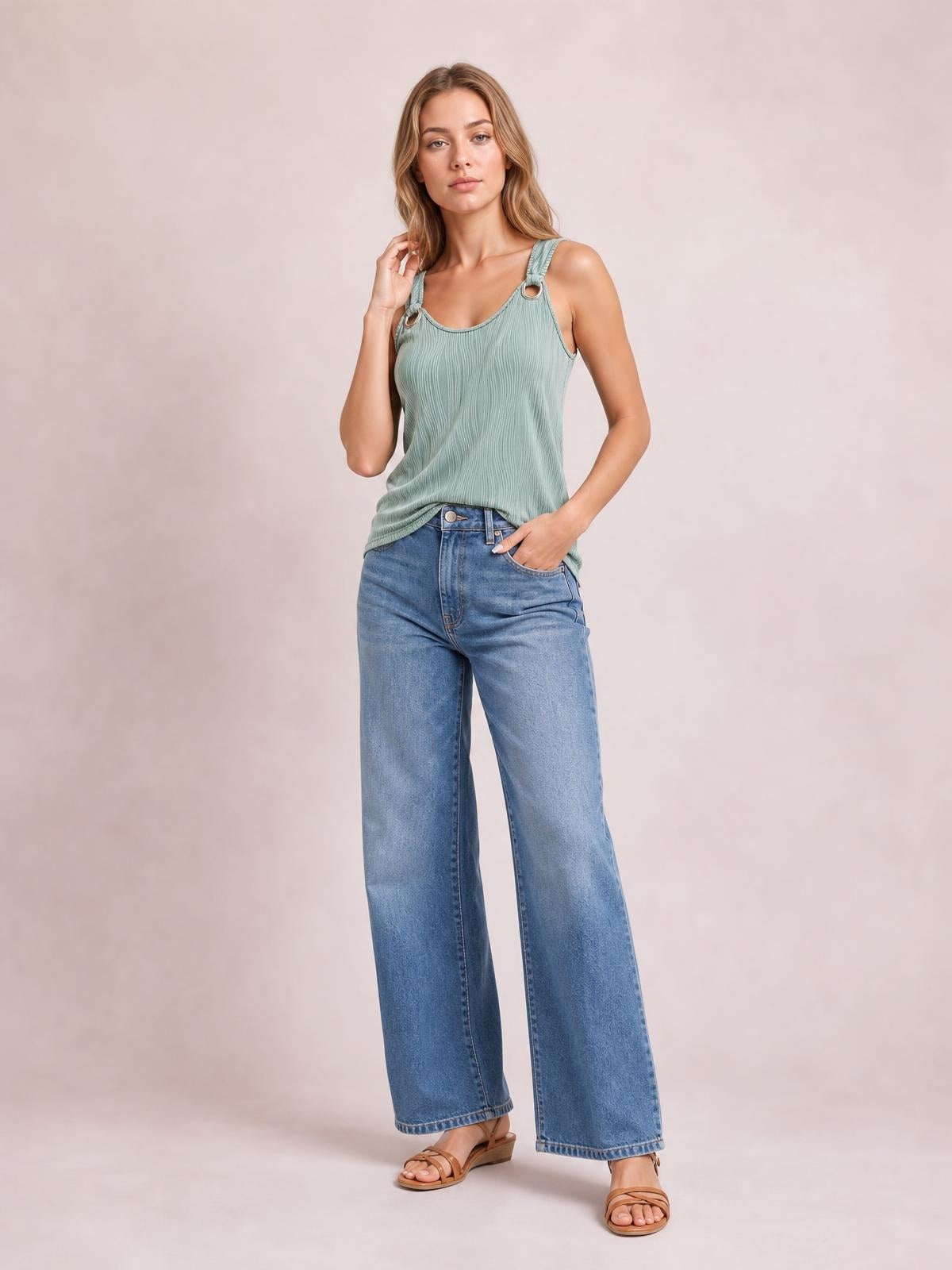A model poses in a sage green round neck wide strap tank with a softly draped, ribbed silhouette and ring-accented straps, one hand in her pocket.