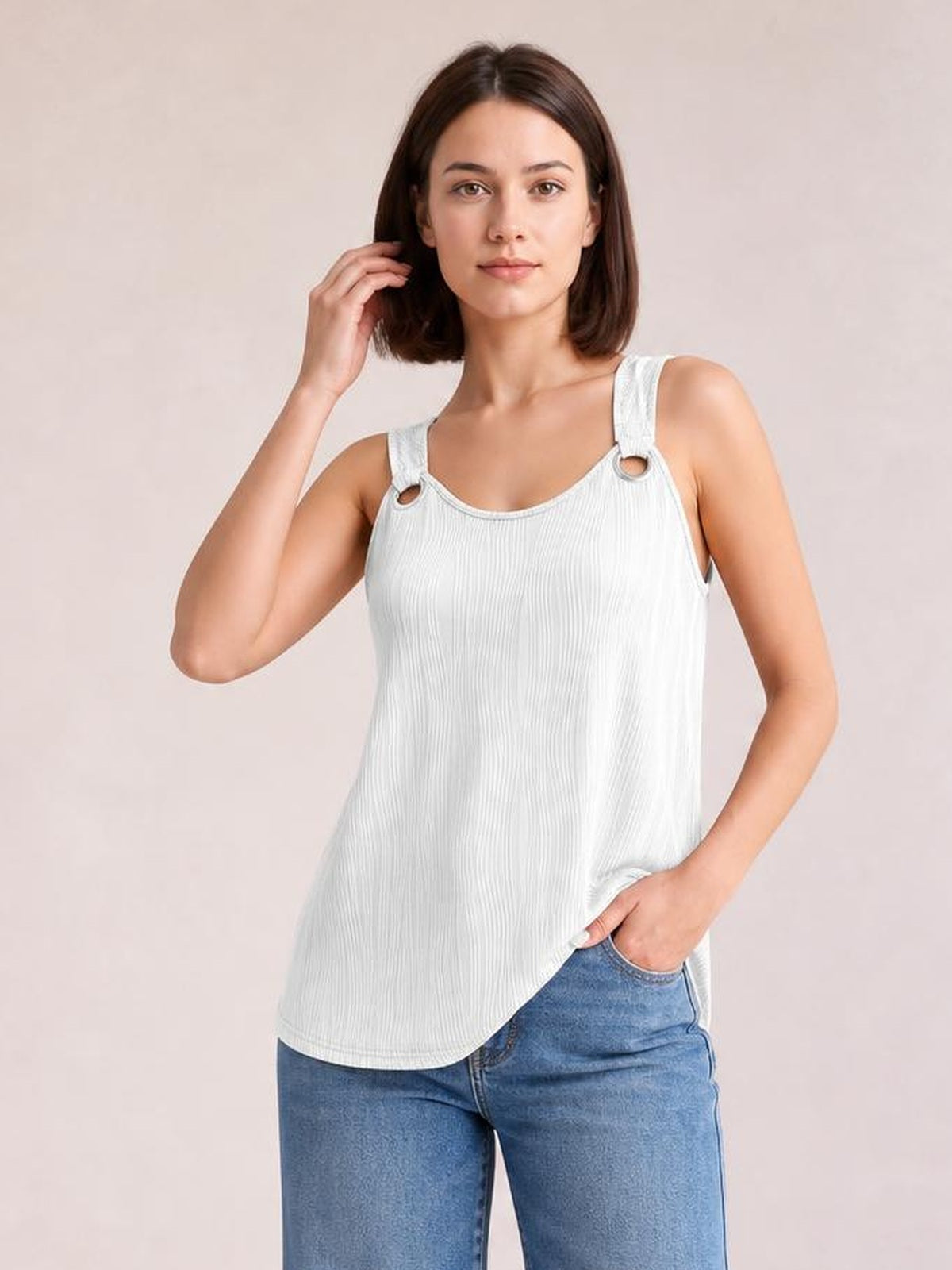A model wears a white round neck wide strap tank with a relaxed, slightly flared silhouette, textured vertical fabric, and a curved hem while posing with one hand in her jeans pocket.