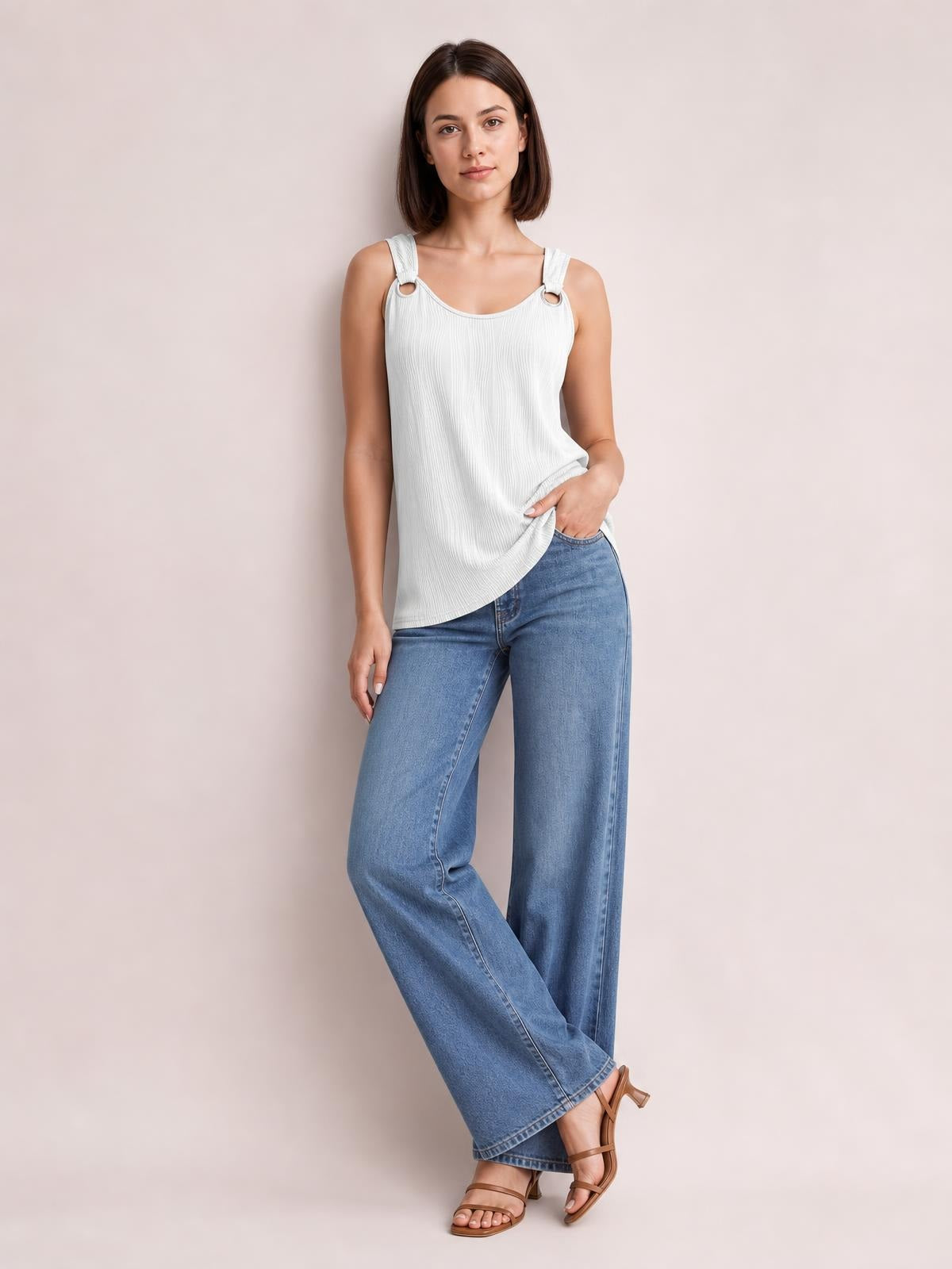 A model wears a white round-neck wide-strap tank with softly pleated texture, relaxed silhouette, and curved hem, posing with one hand at the hem and crossed ankles.