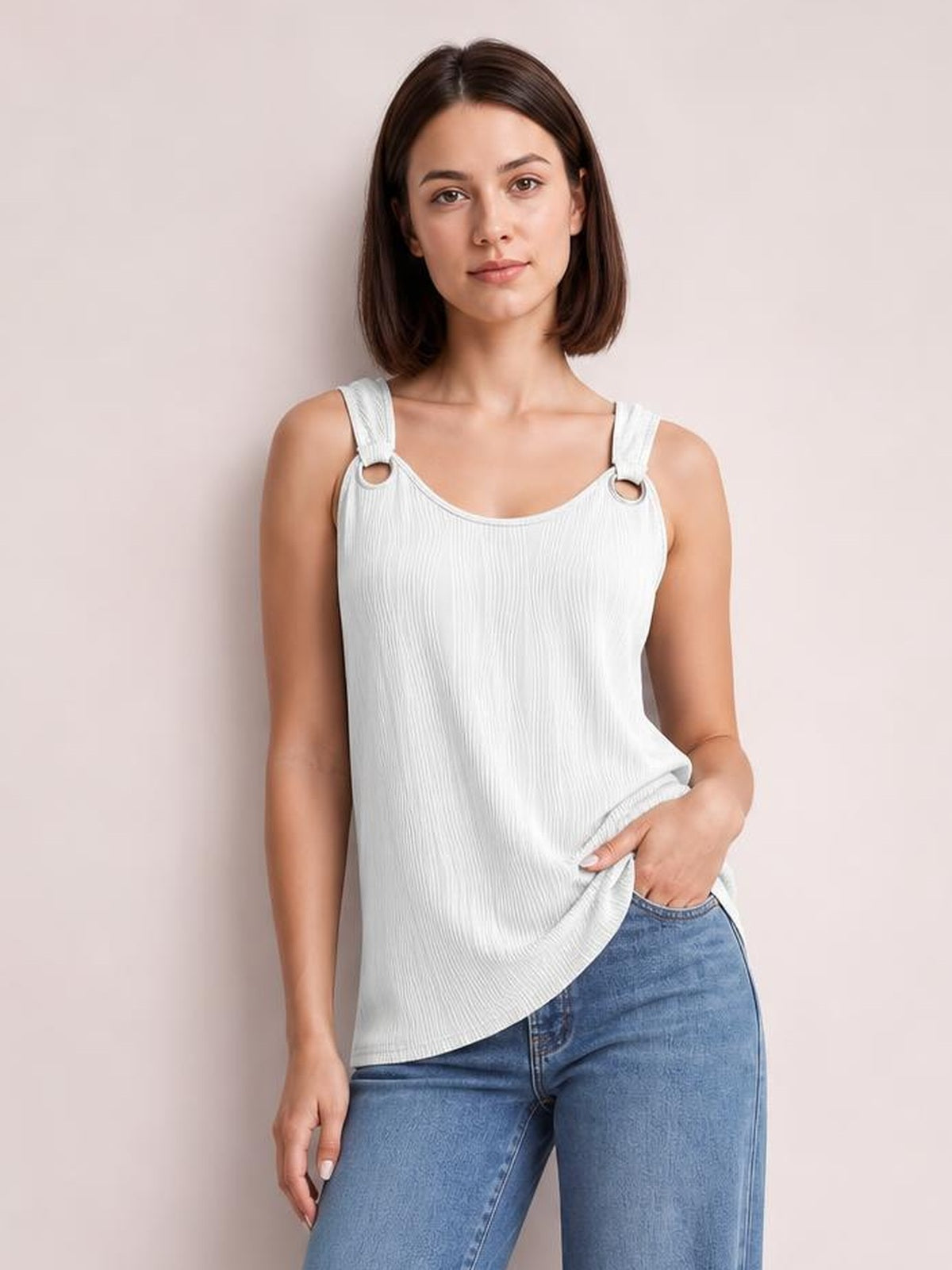 A woman models a white round neck wide strap tank with a relaxed silhouette, textured fabric, ring accents at the straps, and a curved hem over jeans.