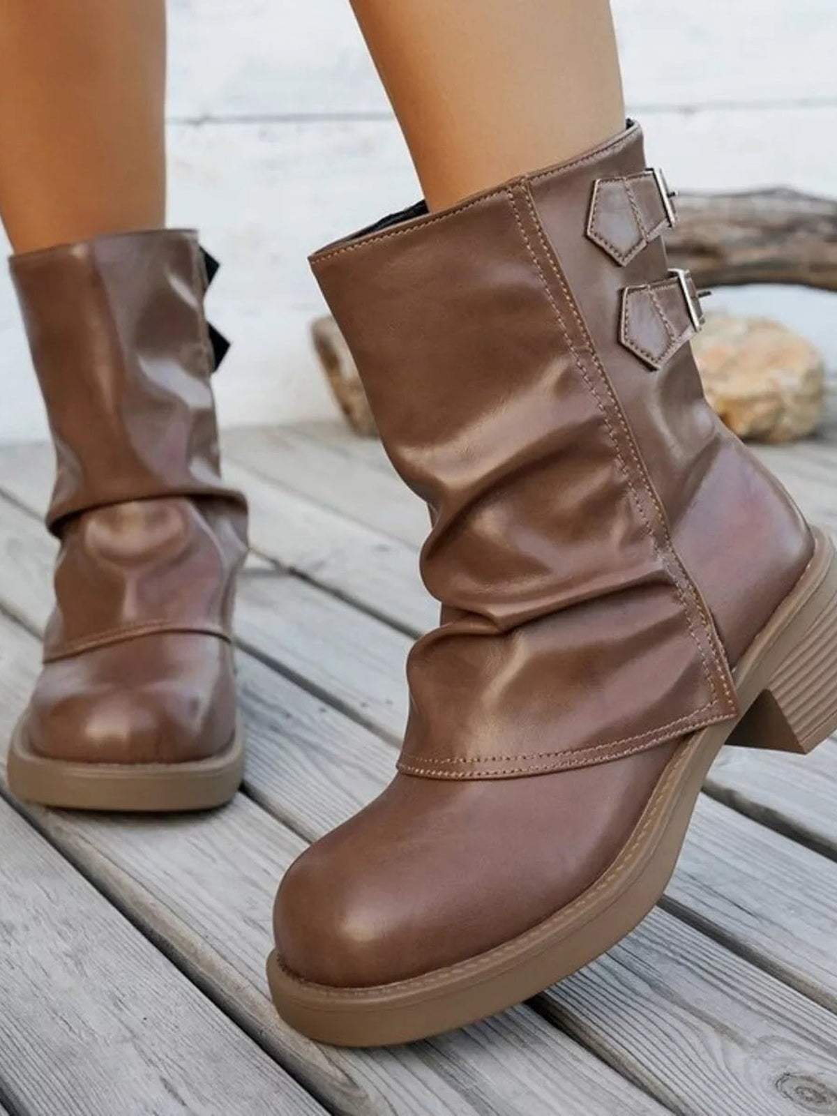 Close-up of brown PU leather round-toe block heel boots with ruched shafts, double buckle accents, stitched panels, and low heels worn by a model.