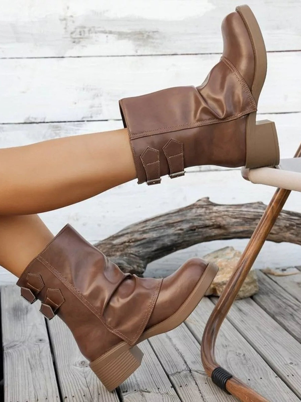 A model crosses her legs while wearing brown PU leather round toe block heel boots with low stacked heels, stitched panels, and decorative ankle straps.