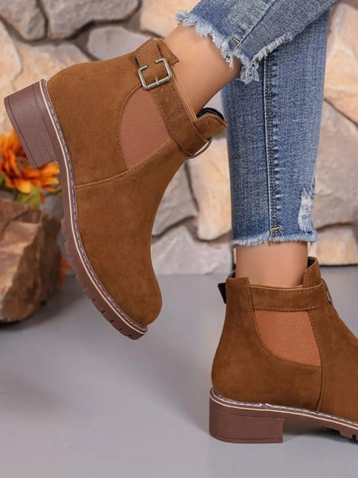 A model wears brown round-toe block heel boots with ankle-high construction, buckle straps, elastic side panels, and sturdy rubber soles with cuffed jeans.