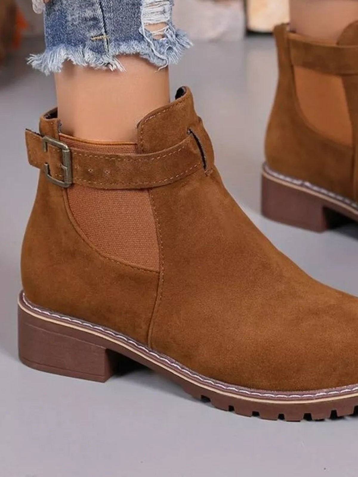 Close-up of brown round-toe block-heel boots with ankle buckle straps, stitched panels, and rugged rubber lug soles, worn with frayed jeans.