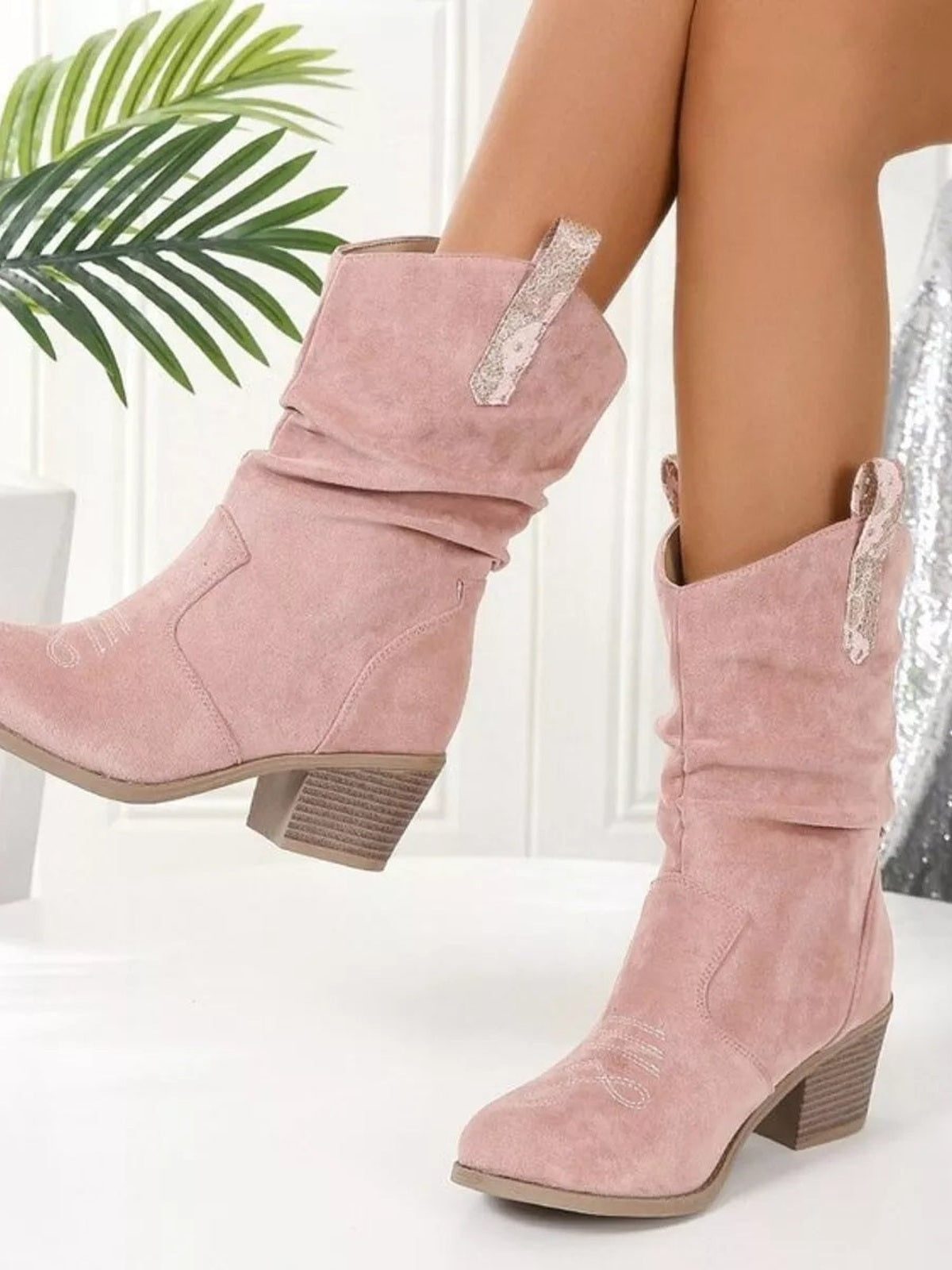 Model displays blush pink round-toe boots with slouchy shafts, stitched panels, decorative side zippers, and low stacked block heels.