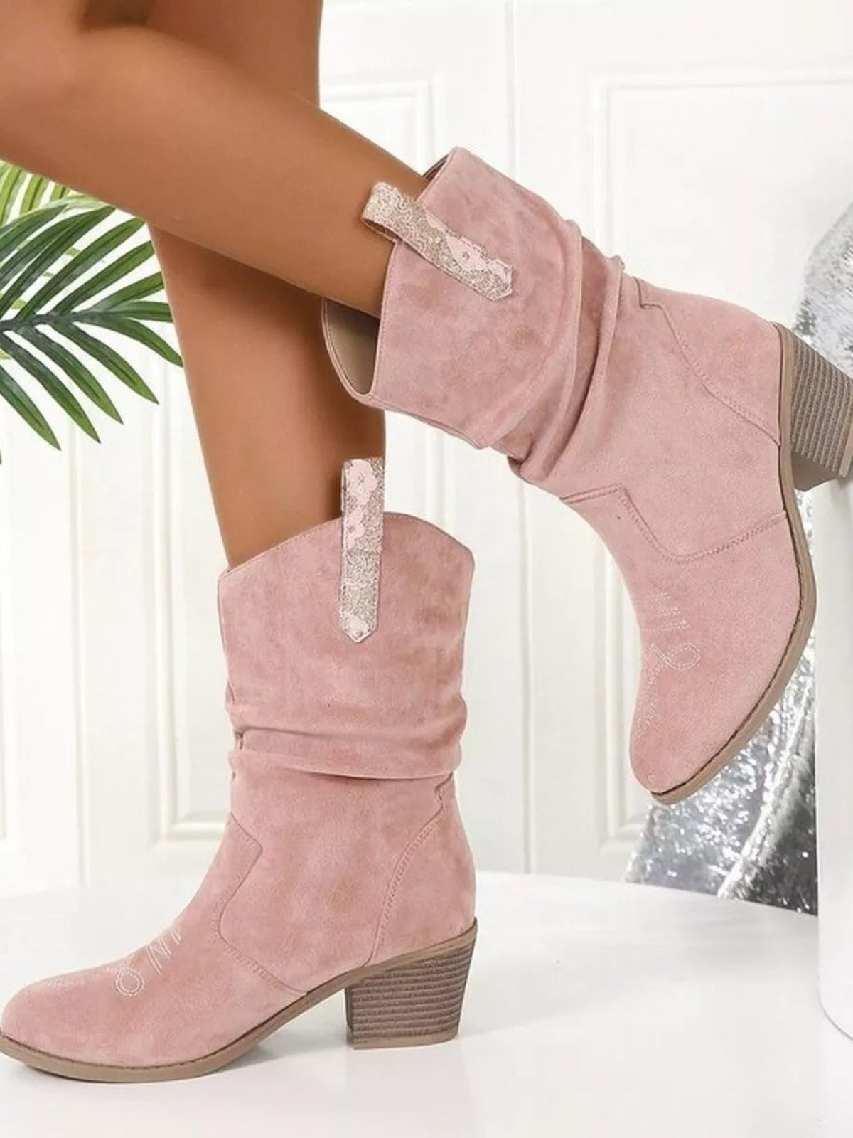 A model poses in pink PU leather boots with round toes, low block heels, slouchy shafts, and decorative metallic side straps.