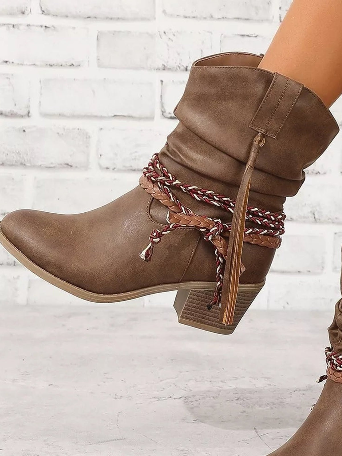 A model wears brown PU leather round toe block heels boots with slouchy shafts, braided wrap detailing, tassel accents, and low block heels.