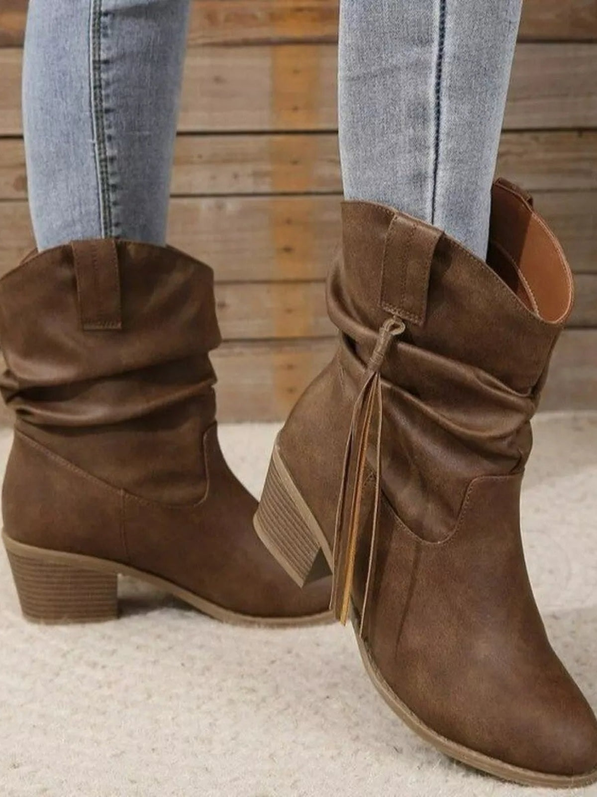 A model wears brown PU leather round-toe boots with slouchy shafts, visible side zippers, low block heels, and one foot posed forward.