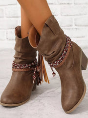 Model poses with crossed legs in brown PU leather round toe block heel boots featuring low heels, ankle shafts, braided wraps, and fringe accents.