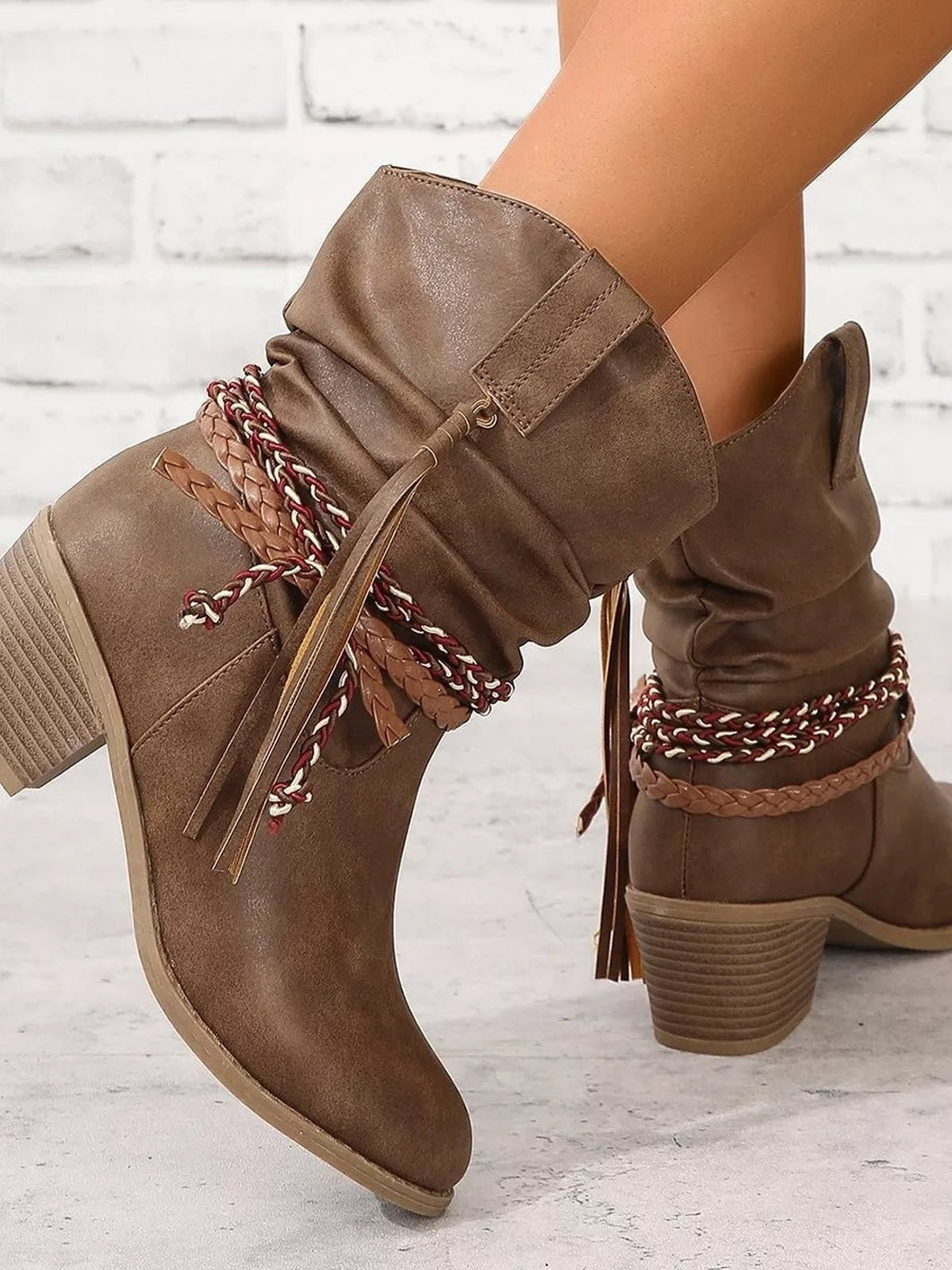 A model poses with crossed legs in brown PU leather boots featuring ruched shafts, round toes, low block heels, braided straps, and tassel accents.