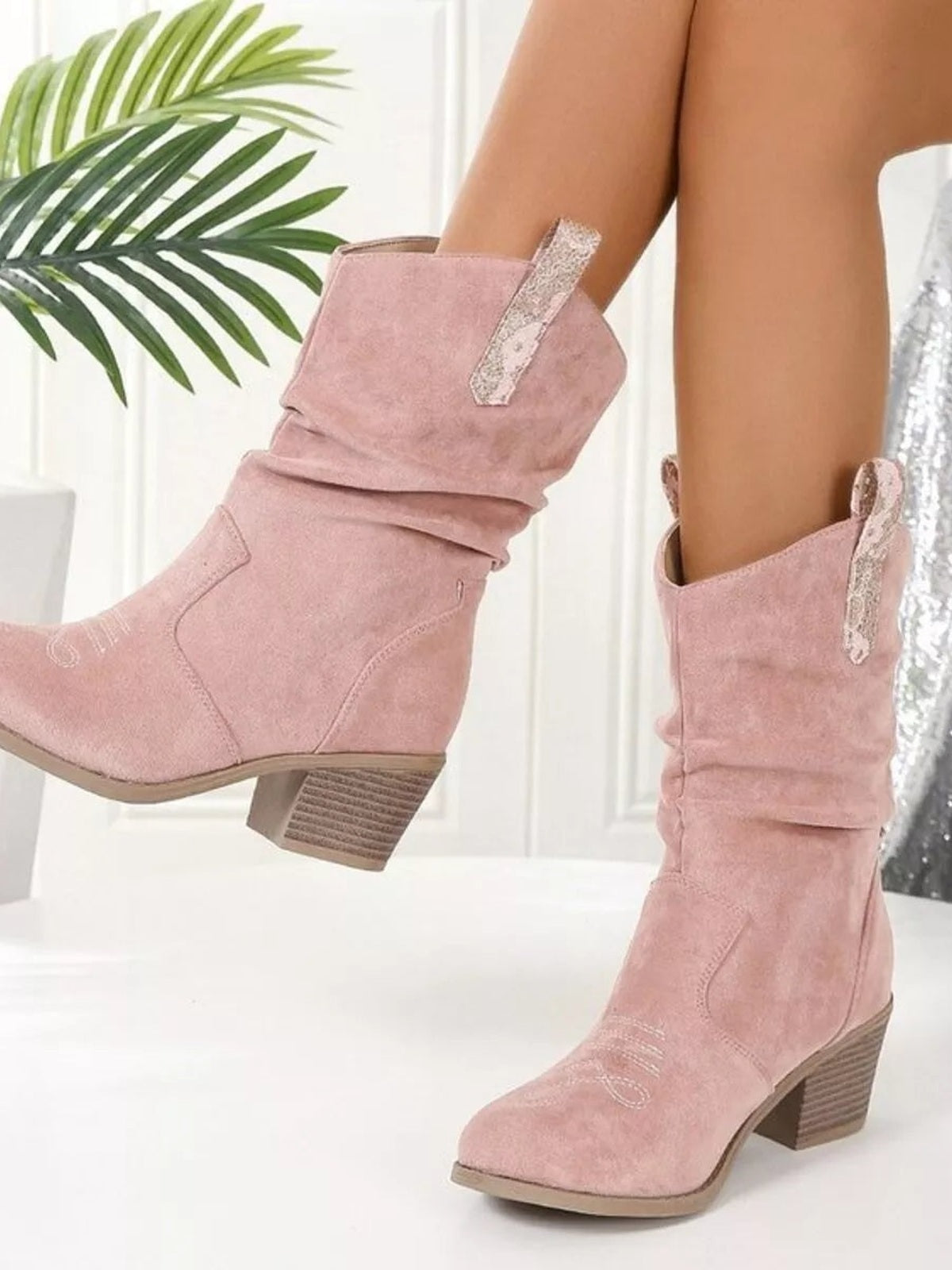 Model displays blush pink round-toe boots with slouchy shafts, stitched panels, decorative side zippers, and low stacked block heels.