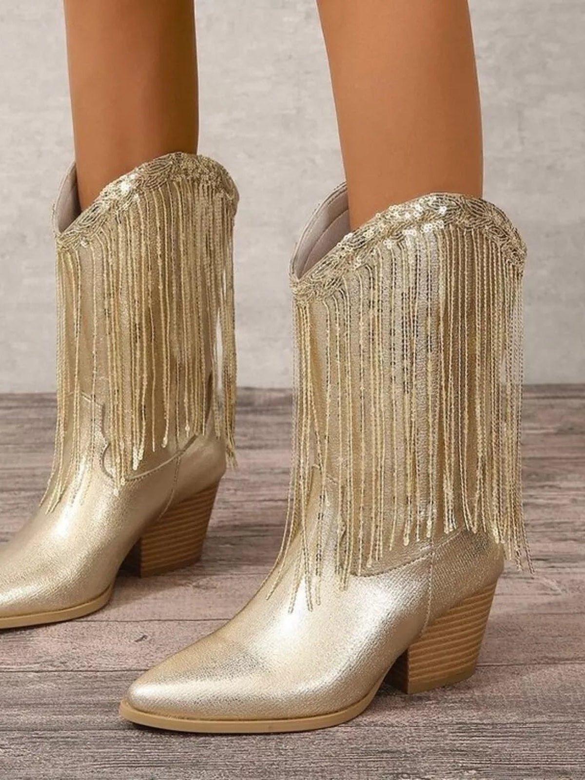 A model wears metallic gold PU leather boots with rounded toes, low block heels, and long shimmering fringe detailing along the shaft.