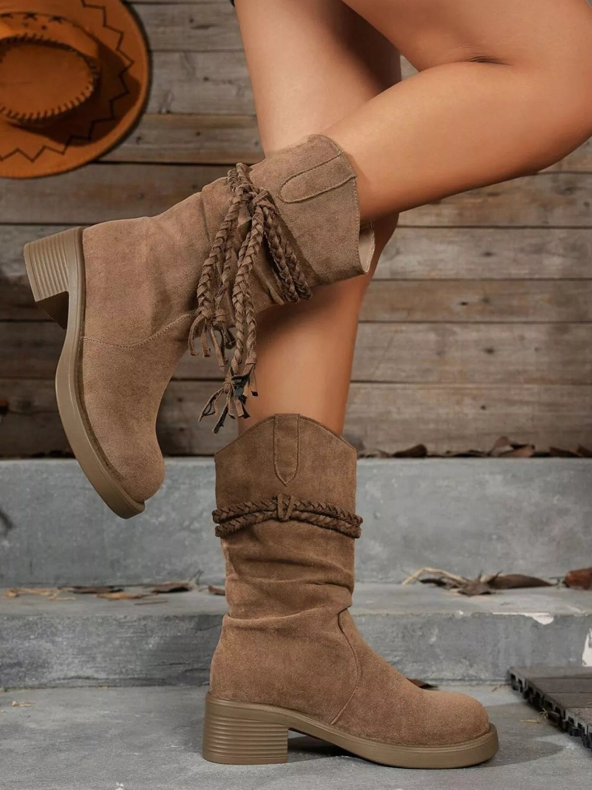 A model poses with crossed legs in taupe round-toe block heel boots featuring mid-height heels, soft slouching shafts, and braided wraparound ties.