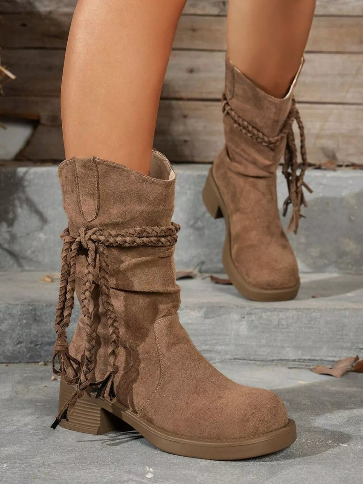 Model wears taupe round-toe block heel boots with a mid-height heel, slouchy shaft, and braided wraparound lace detailing in PU leather.