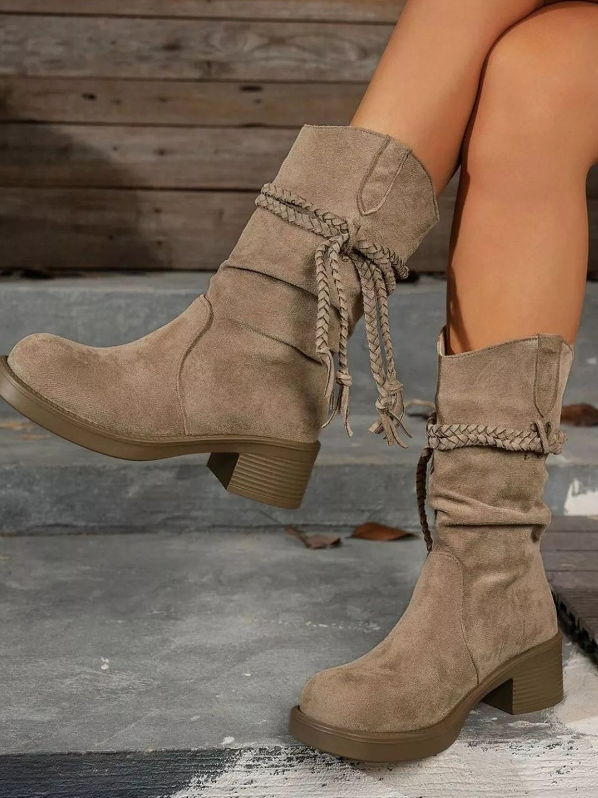 A model wears taupe round-toe block-heel boots with cuffed shafts and braided wraparound ties, lifting one foot to show the mid heel and textured sole.