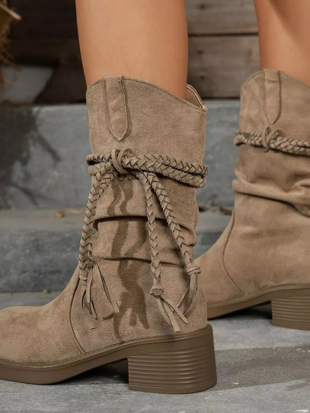 A model wears tan round-toe block heel boots with mid-height stacked heels, braided wraparound ties, fringe accents, and stitched PU leather construction.