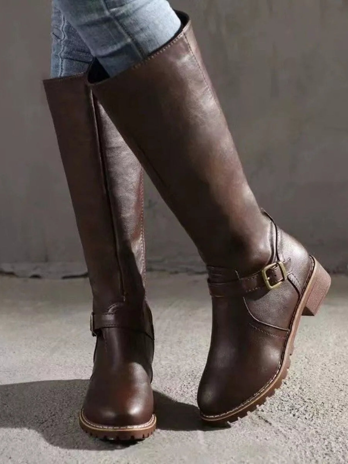 A model crosses her legs in dark brown PU leather boots with tall shafts, round toes, buckle straps, low block heels, and textured rubber soles.