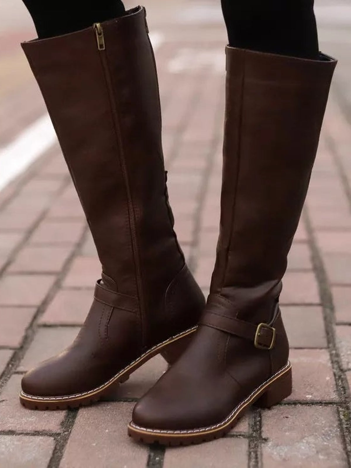 A model wears dark brown round-toe boots with tall shafts, side zippers, ankle buckle straps, stitched trim, and low block heels.