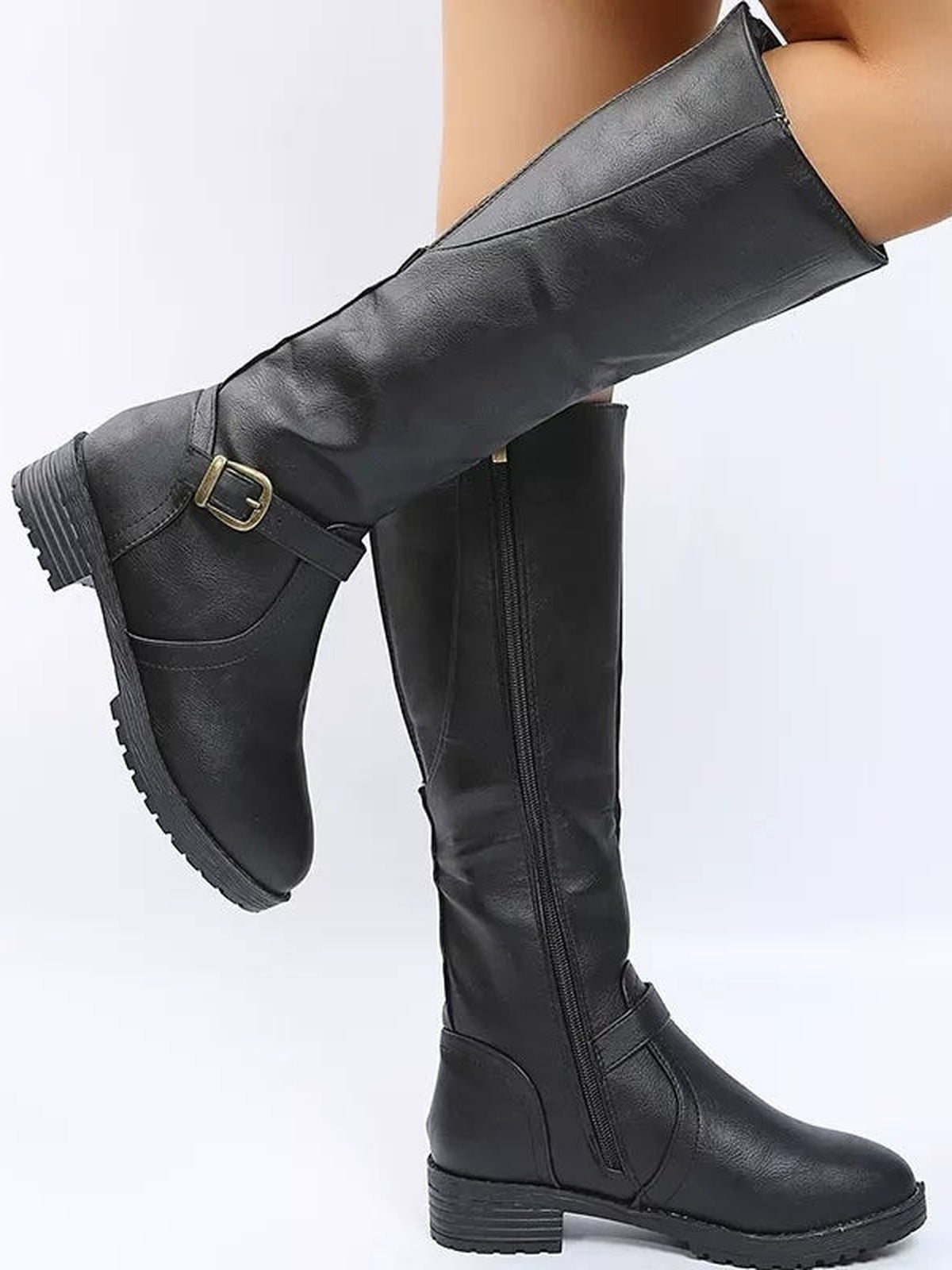 A model wears black PU leather knee-high boots with round toes, low block heels, side zippers, buckle straps, and rugged rubber soles, one leg lifted.