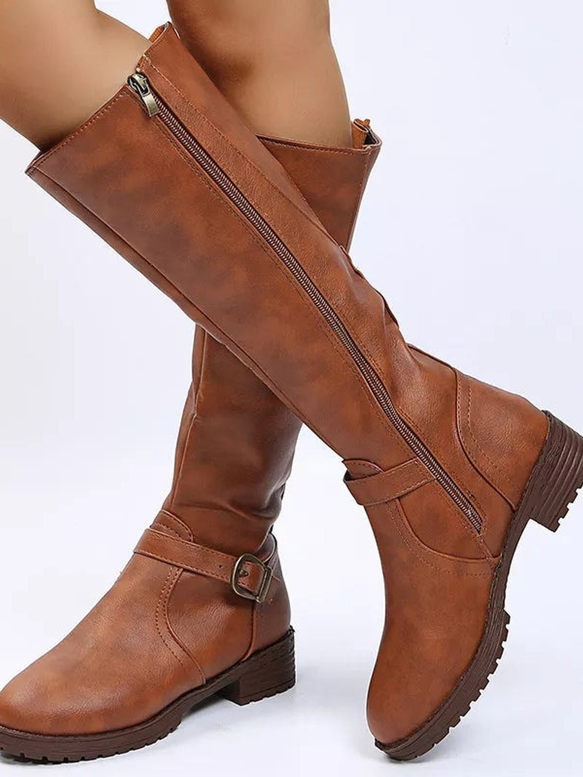 A model wears brown PU leather boots with round toes, low block heels, side zippers, buckle straps, and rugged rubber soles in a crossed-leg pose.