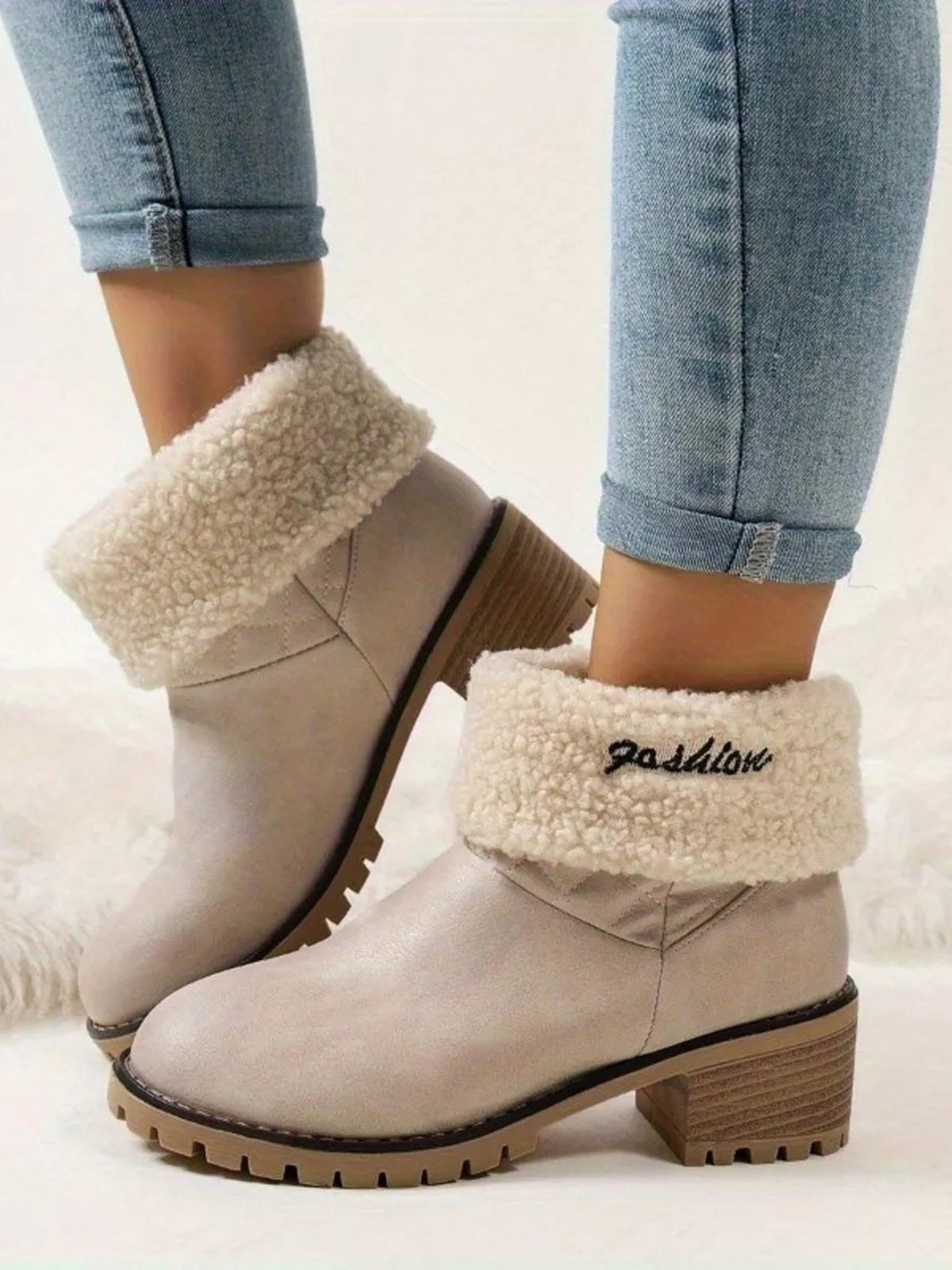 A model wears beige round-toe boots with plush fleece cuffs, low block heels, stitched panels, and chunky lugged rubber soles over cuffed jeans.